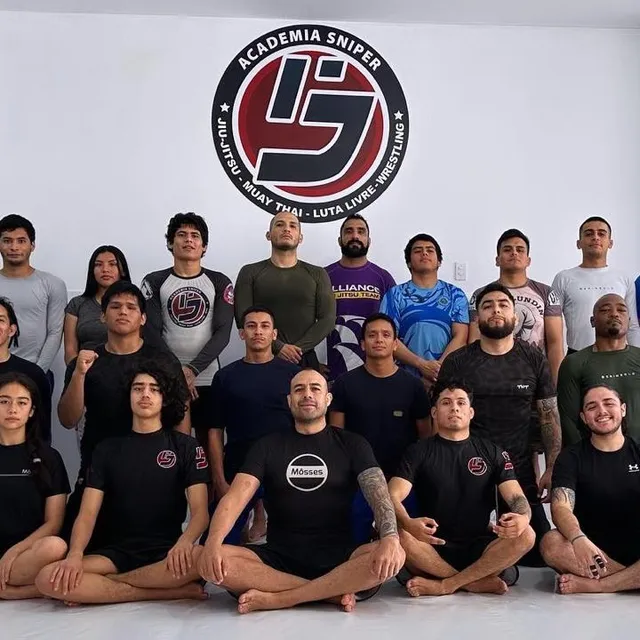 Grupo de alumnos con ropa casual y gi posando de pie frente al logo de la academia