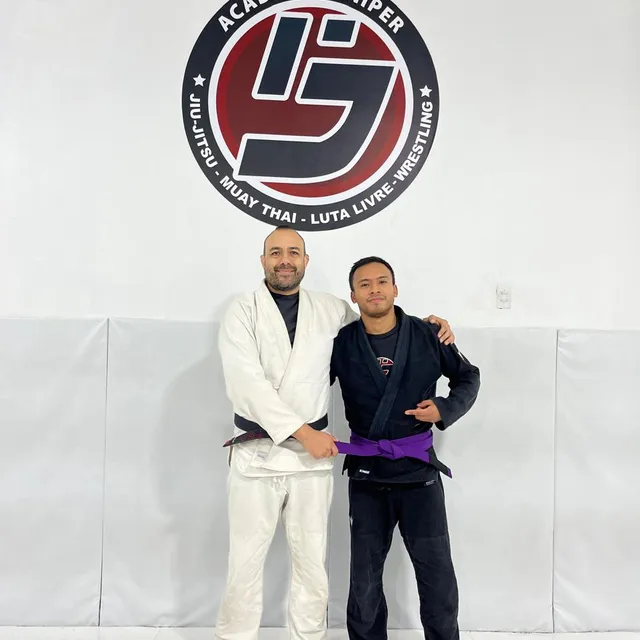 Instructor posando con un alumno de cinturón morado tras una entrega de grado, frente al logo del gimnasio